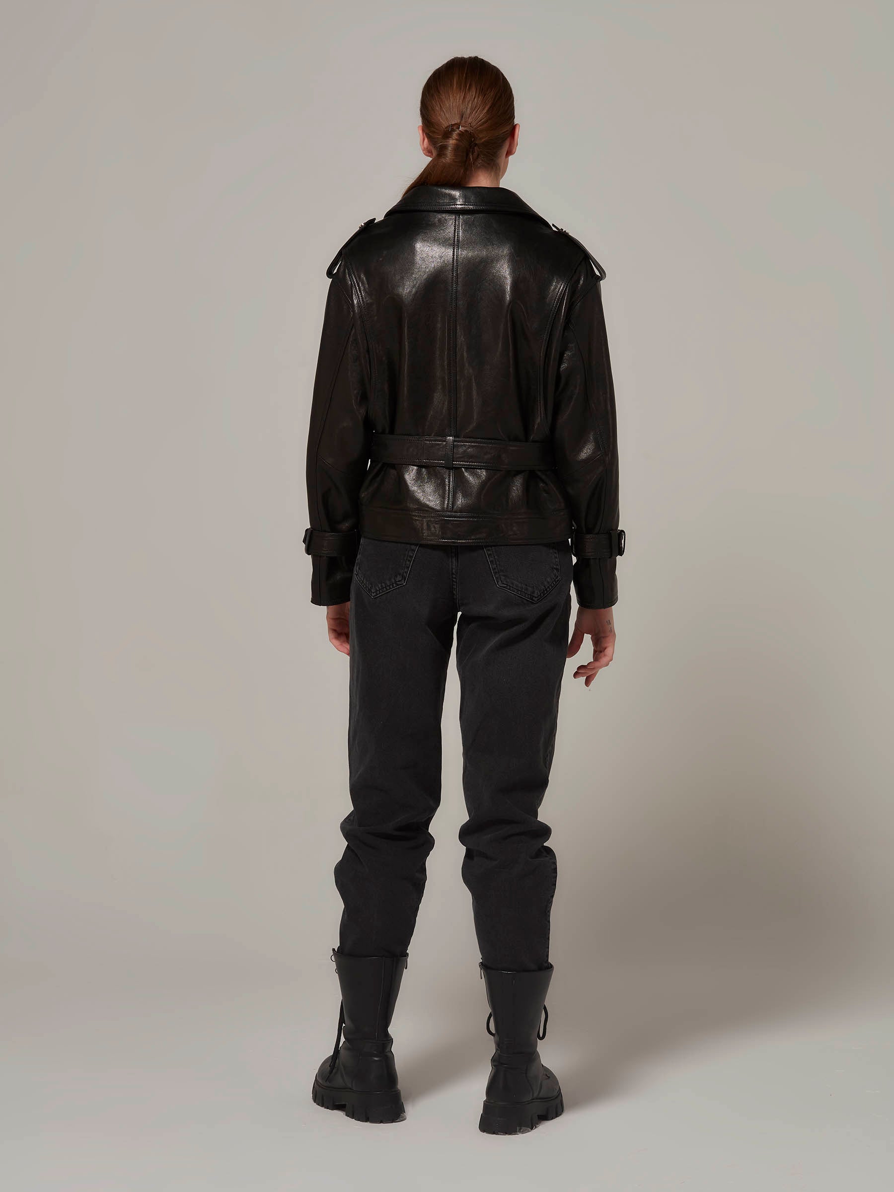 Woman in black biker leather jacket with belt, rebellious attire, motorcycle jacket, moto style, standing. (back view)