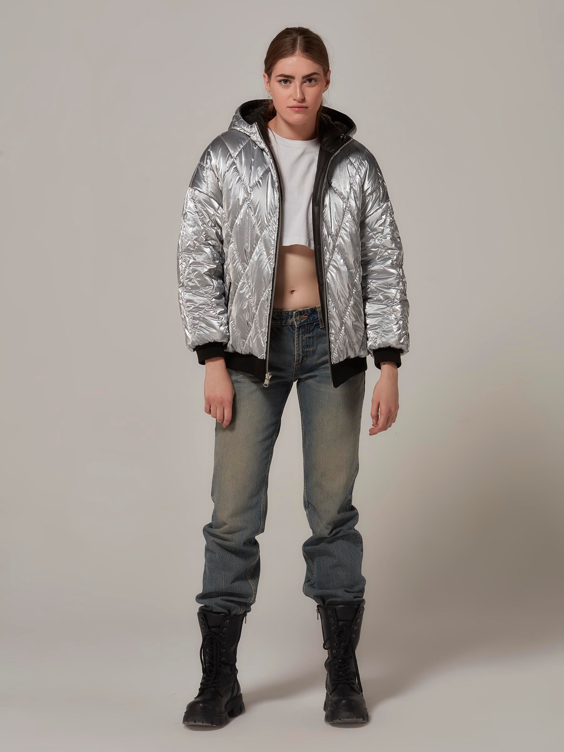 A rebellious-looking woman stands in a front view, wearing a black lambskin leather and metallic silver waterproof fabric double-sided reversible puffer jacket. Full length photo.