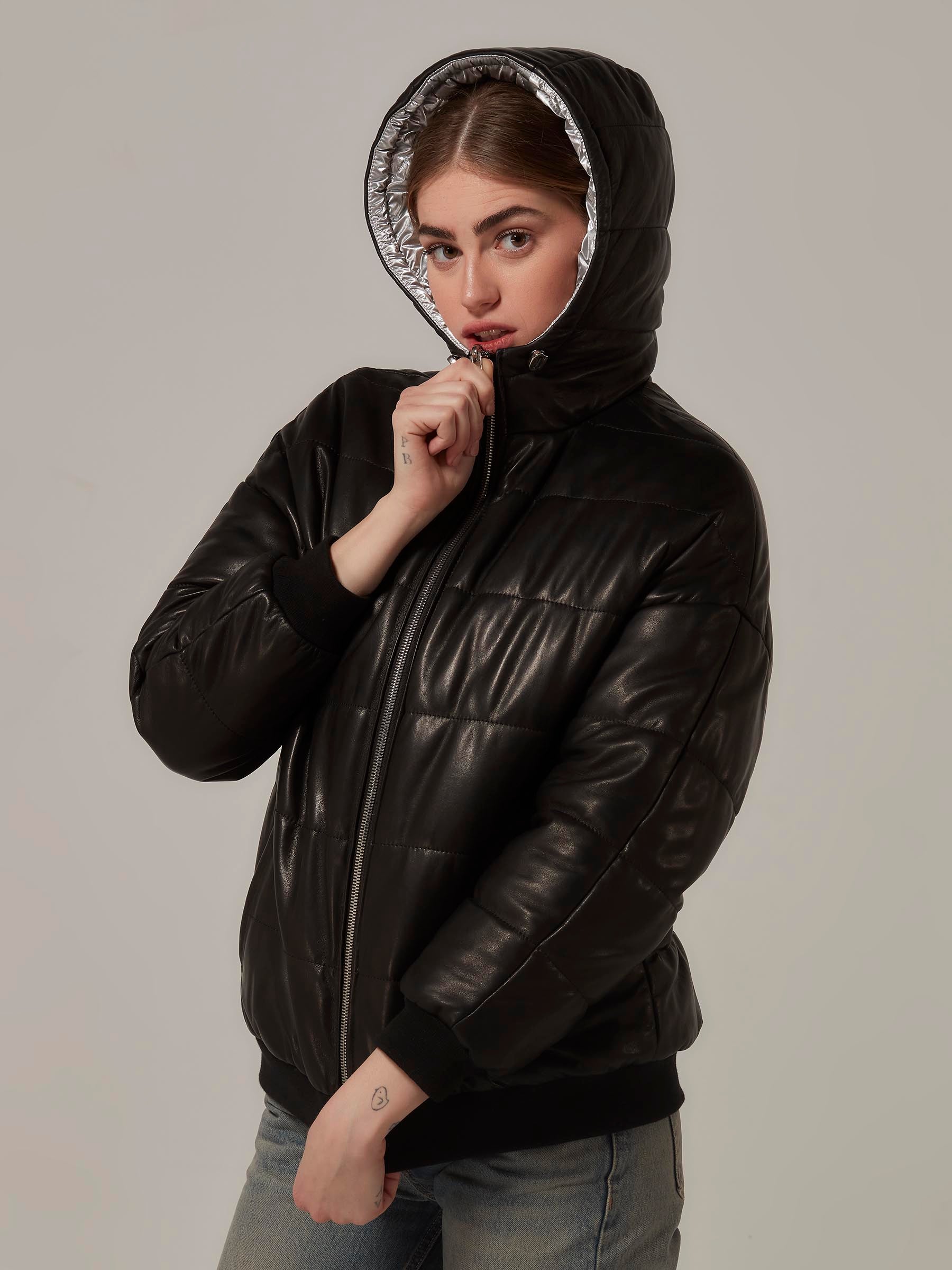 A stylish woman in a black leather and metallic silver reversible puffer jacket poses rebelliously. Close-up, three-quarters front view, as she coses the front zipper and adjusts the hood, exuding a fashionable vibe.