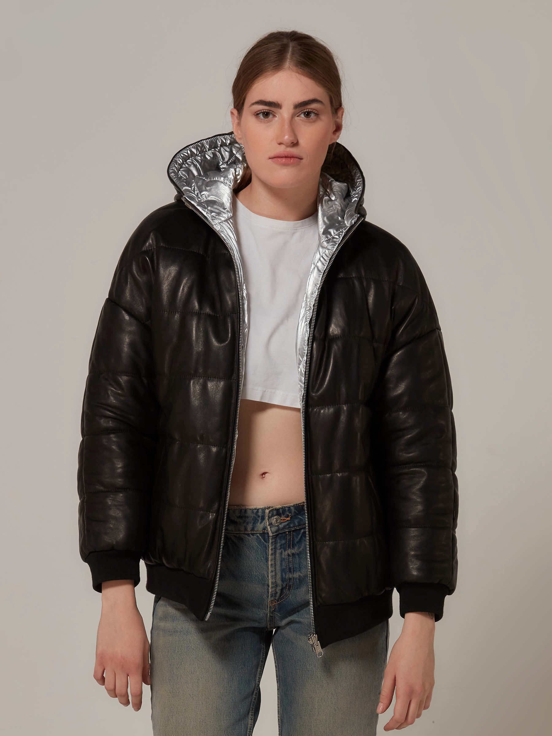 A rebellious-looking woman stands in a front view, wearing a black lambskin leather and metallic silver waterproof fabric double-sided reversible puffer jacket. Three quarters close up photo.