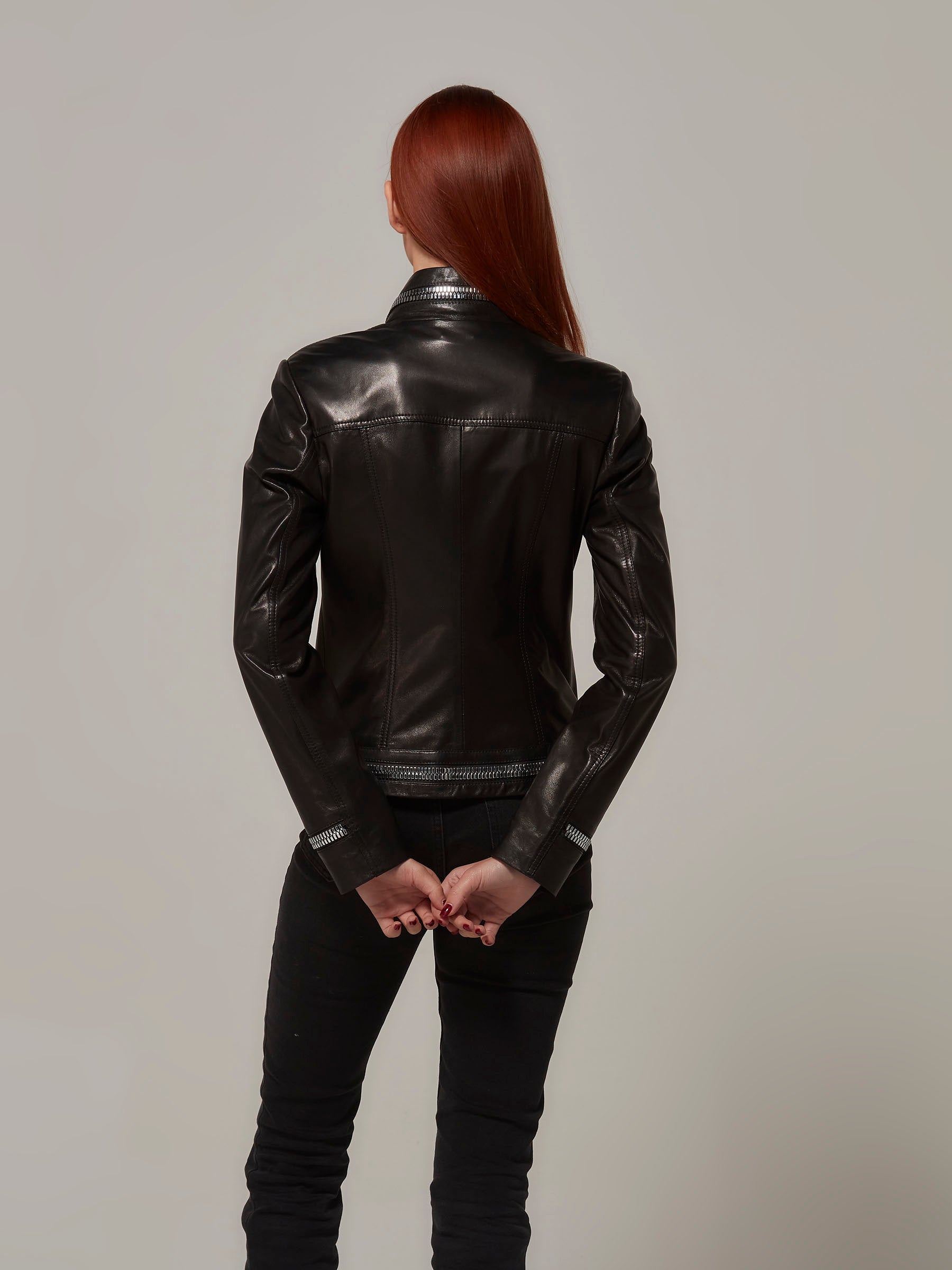 Back view of a chic woman wearing a Black biker jacket with metal zipper details on collar, sleeves, and hem. Three quarters length photo.