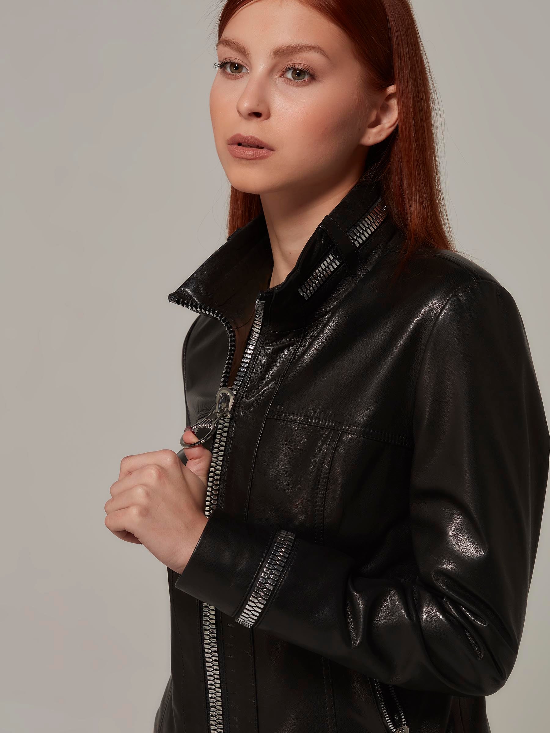 Close-up photo of a fashionable woman wearing a black biker jacket with metal zipper details on the collar and sleeves. She holds the zipper head for a glamorous pose.