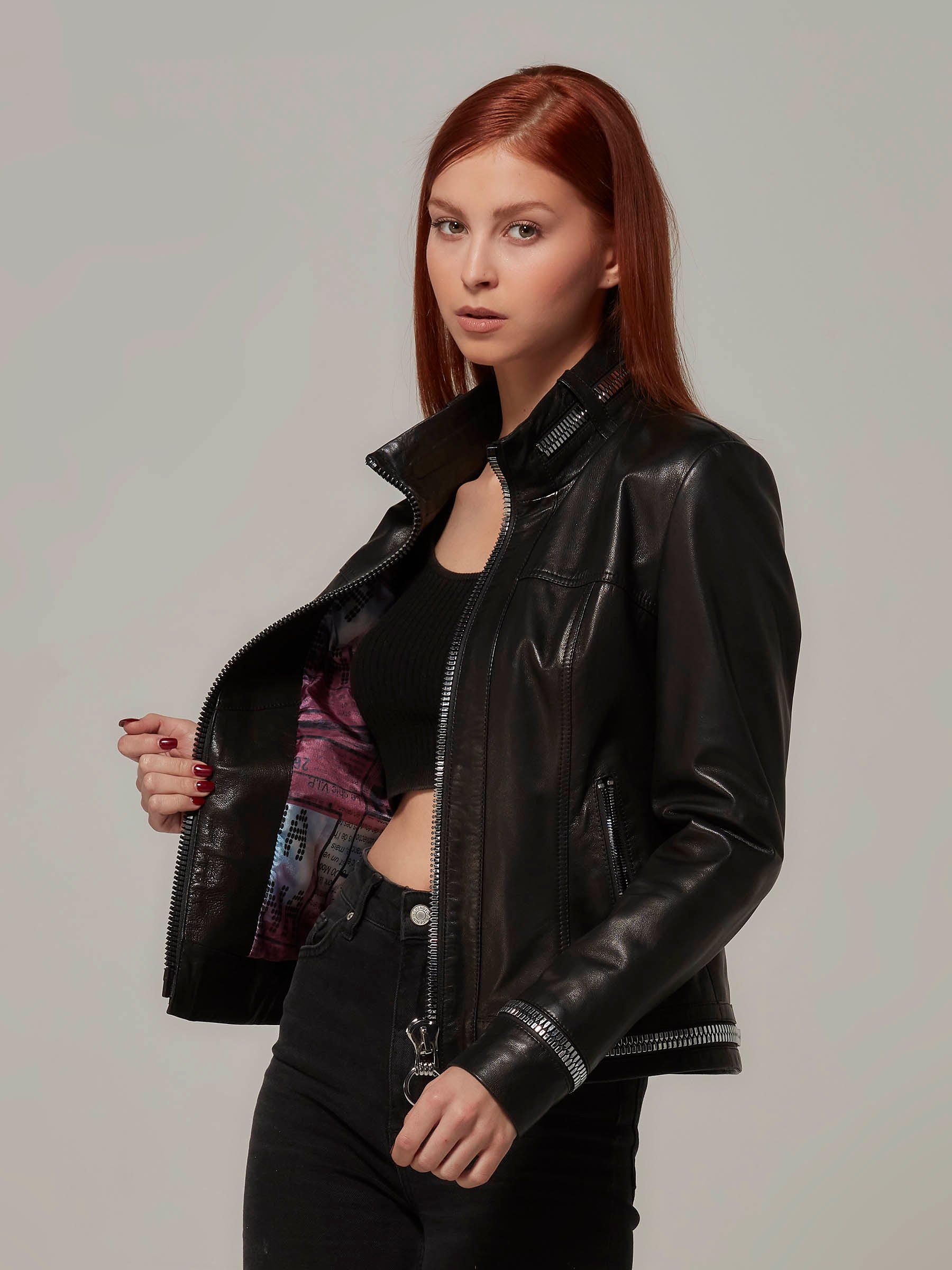 A stylish woman in a black biker jacket with metal zipper details on the collar and sleeves. She is holding the right side of the jacket to reveal the inner side. Close-up photo.