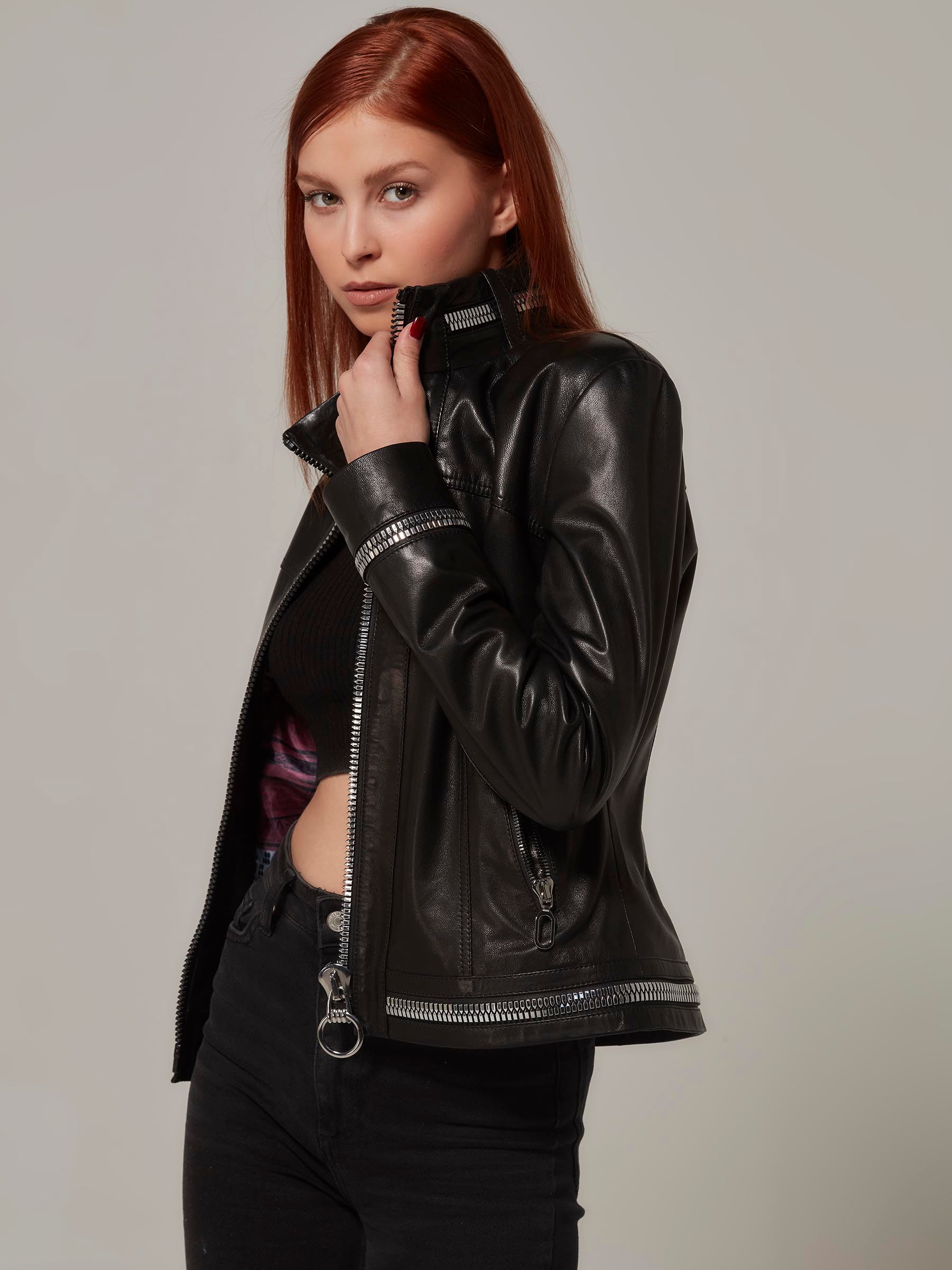 Close-up front view photo of a chic woman in a black biker jacket. She holds the left side of the collar, showcasing the metal zipper details. Her pose exudes charm and style.