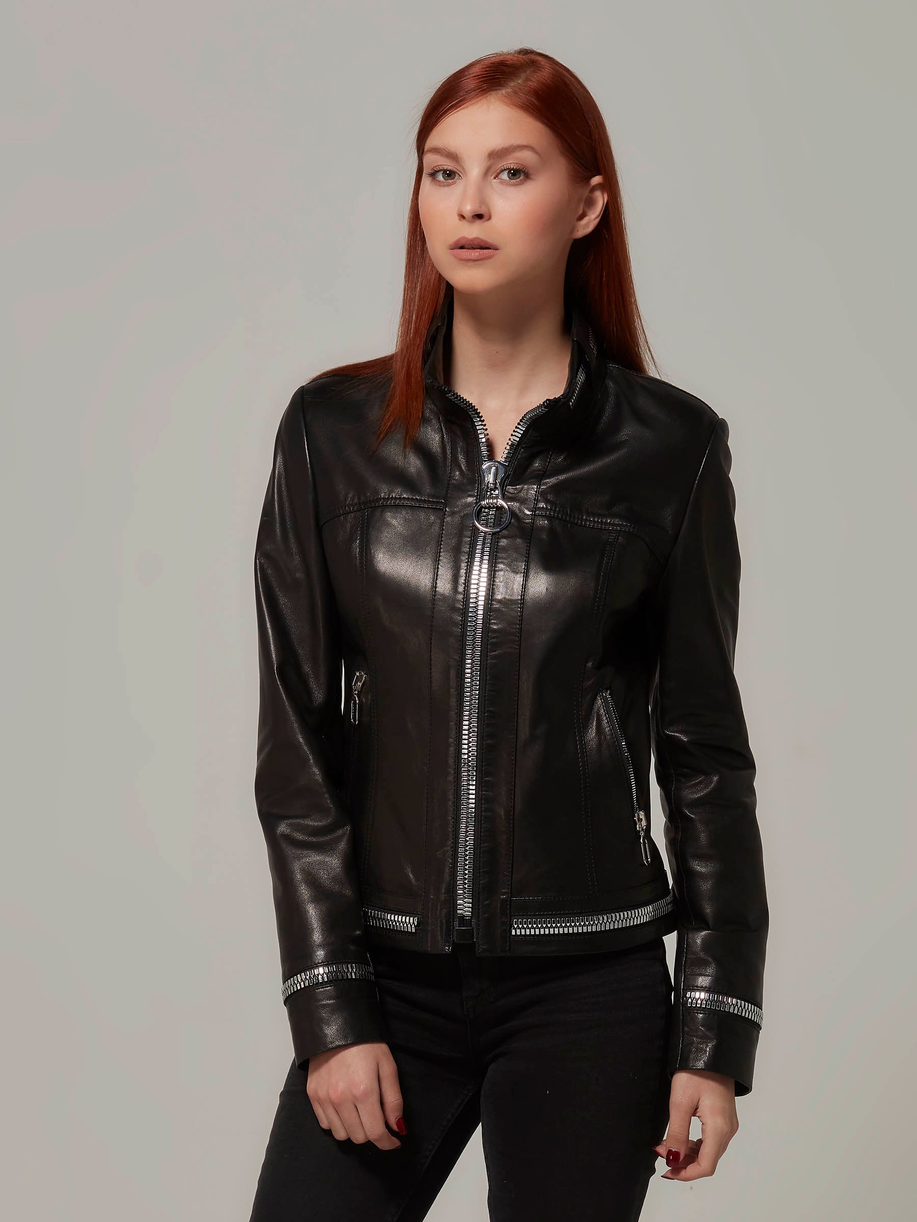 Front view of a chic woman wearing a Black biker jacket with metal zipper details on collar, sleeves, and hem. Three quarters length photo.