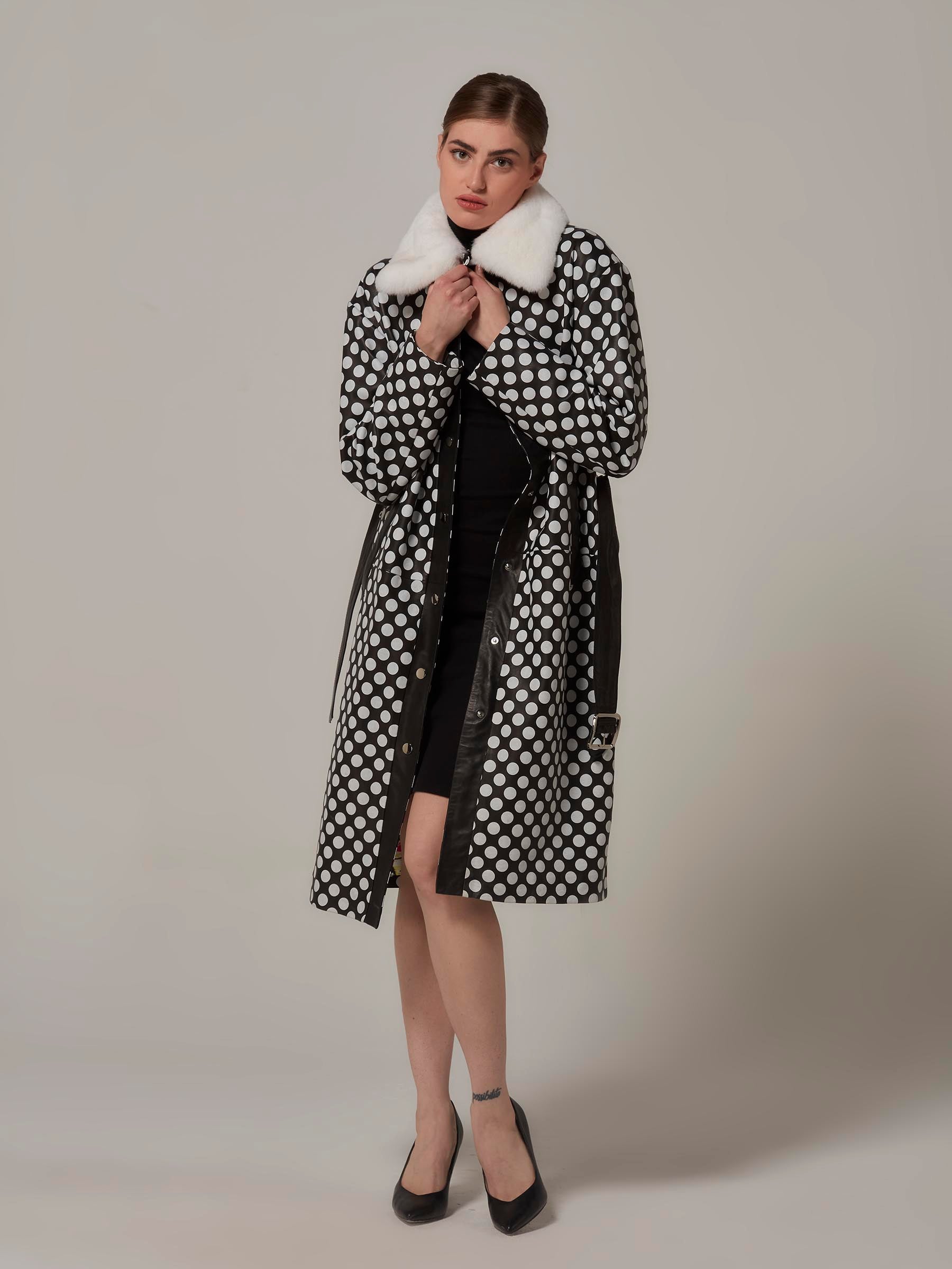 A stylish woman poses front view in a Black and White Leather Coat with Polka Dot, charmingly holding the rex fur collar from both sides. Full-length photo.