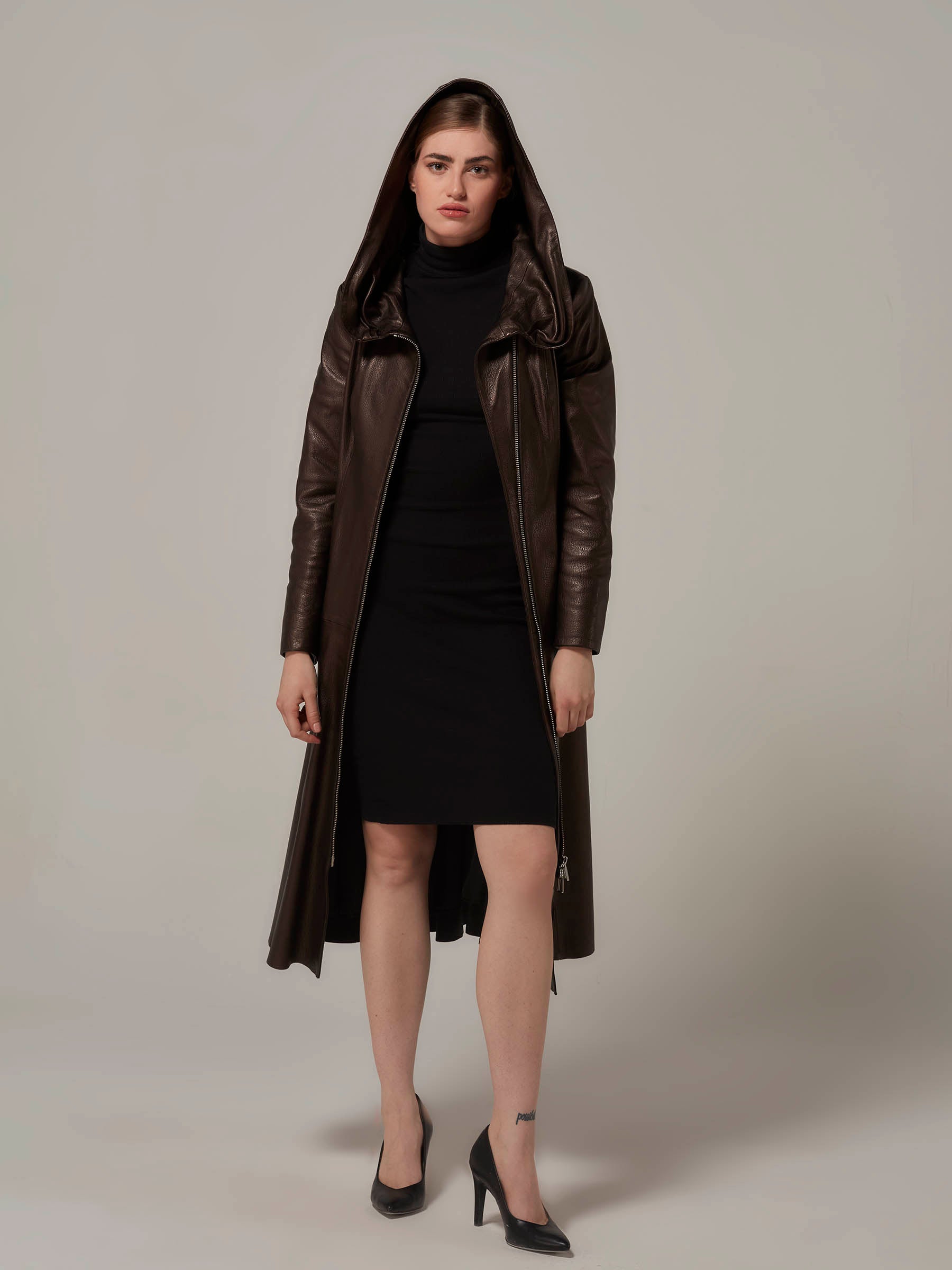 A beautiful woman in a black dress and leather coat, captured in a full-length photo. She is donning a fashionable dark bronze color lambskin hooded leather coat with the hood up and front zipper open, confidently posing in a front view.