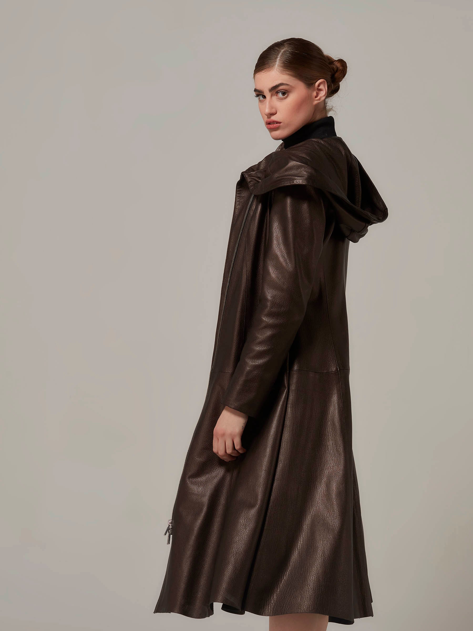 In a three-quarters length photo, a stunning woman strikes a striking pose while wearing a dark bronze lambskin hooded leather coat in a side view.