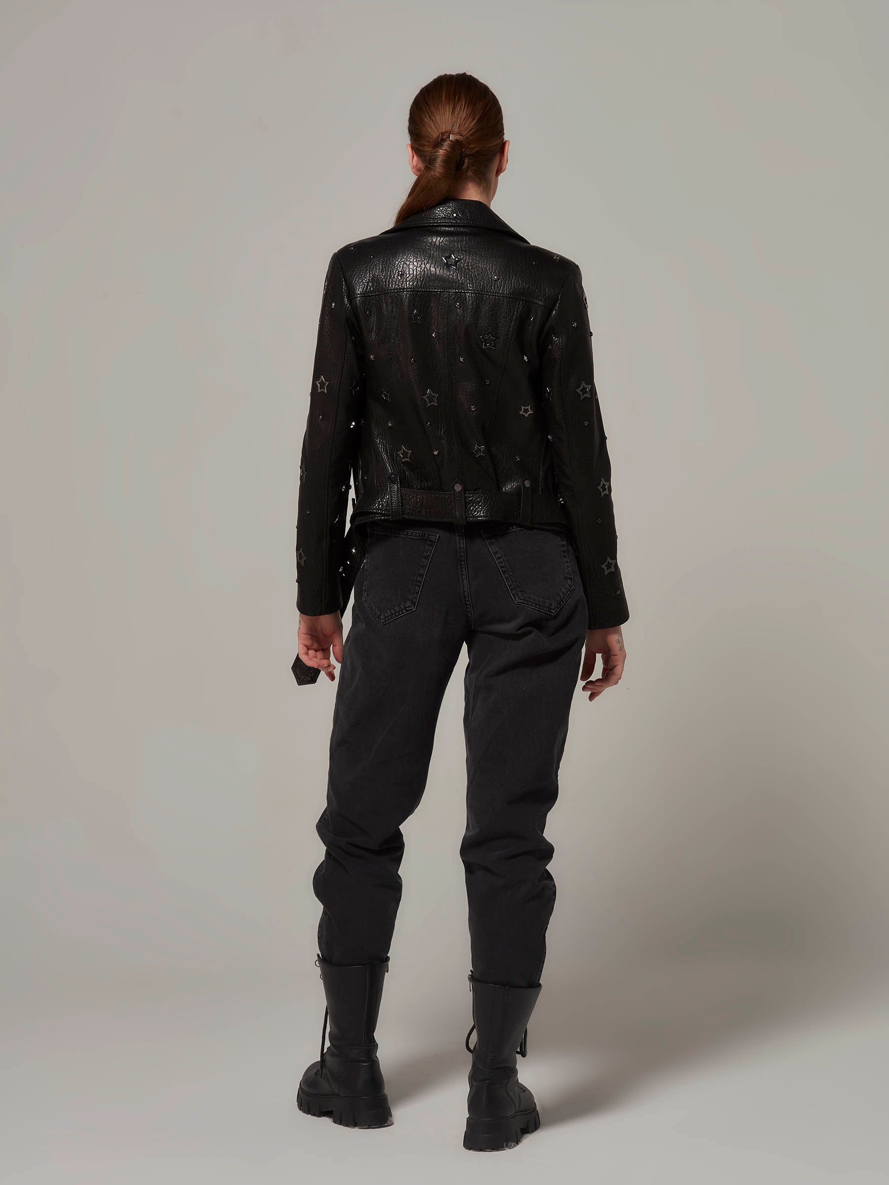 Woman in a black, biker jacket adorned with star details, with the unique texture of the leather and embodying a rebellious rock attitude. Standing back view pose.