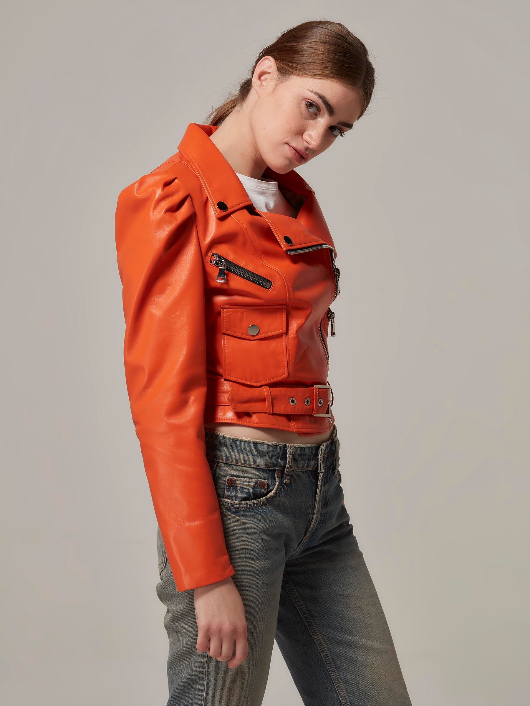 Person wearing an orange leather jacket and gray jeans on a plain background